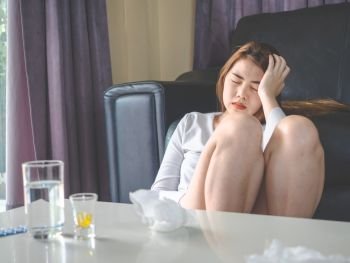 sickness and gynecology concept from woman with allergy, headache and fever sitting at sofa in home, have pills and glass of water laying the table.
