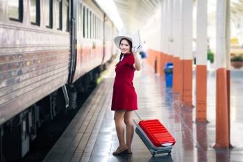 Asian woman pregnant in red dress holding a map and hand up say hello with red suitcase in railway station travel.