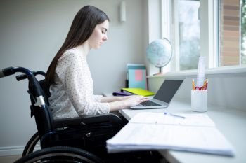 Teenage Girl In Wheelchair Studying At Home On Laptop