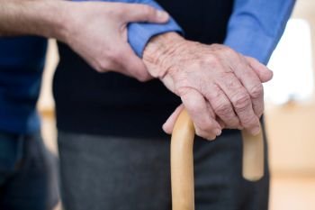 Senior Man’s Hands On Walking Stick With Care Worker In Background