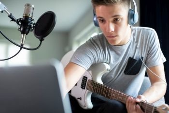 Teenage Boy Playing Guitar And Recording Music Onto Laptop At Home