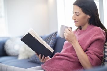 Woman With Hot Drink Relaxing On Sofa At Home Reading Book