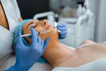 Cosmetician in gloves gives chin botox injection to female patient on treatment table. Rejuvenation procedure in beautician salon. Doctor with syringe and woman, cosmetic surgery against wrinkles. Cosmetician gives chin botox injection to patient