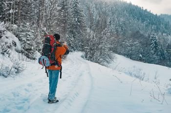 Rear view of tourist taking photo of mountain forest landscape on snowy winter day. Blue jeans, orange garment, red backpack. Hiking travel extreme concept