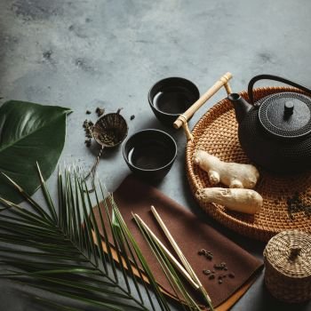 Traditional Asian tea ceremony arrangement. Iron teapot, cups, dried green tea leaves, ginger and tropical leaves over dark concrete background, top view, copy space. Traditional Asian tea ceremony arrangement, top view