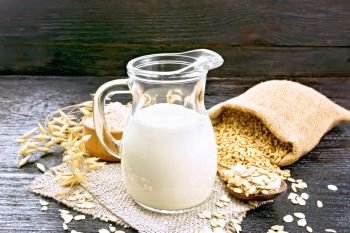 Oat milk in jug, flour in bowl, oatmeal in spoon, grain in jute bag, oaten stalks on burlap against wooden board