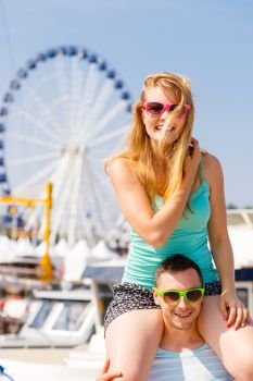 Relationship goals, summer love concept. Man giving girlfriend piggyback ride on marina with ferrish wheel in background.. Man giving girlfriend piggyback ride on marina