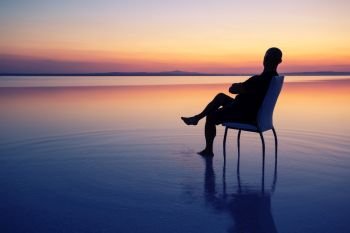 Silhouette of a man sitting on a chair in the middle of the lake at sunset. Calm water, dramatic sunset. concept of privacy, freedom and recreation. Silhouette of man sitting on chair in middle of lake at sunset
