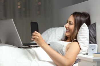 Happy woman using a smart phone and laptop lying on a bed in the night at home