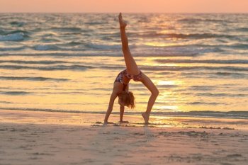 Sweet girl doing yoga on the beach over sunset sky background. Active sportive summer camp. Dancing outdoors. Healthy childhood.. Teen girl doing yoga on the beach