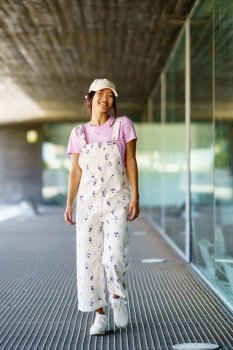 Full body happy young Asian female in stylish clothes and cap, smiling and looking at camera while walking near glass wall of contemporary building in daytime. Cheerful Asian woman outside modern building