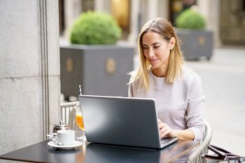 Positive adult female freelancer with blond hair in casual clothes typing on laptop, while working remotely at table with coffee cup and juice glass in outdoor cafe. Smiling self employed woman using laptop in cafe