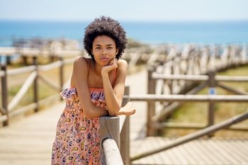 Young African American female tourist in ornamental dress with curly hair, leaning on hand and looking at camera while relaxing on boardwalk against sea and blue sky on summer day on resort. Black woman resting on wooden path near beach