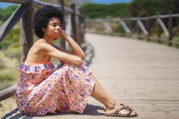 Side view of pensive ethnic female in summer dress and sandals with curly hair, leaning on hand and looking away while sitting on wooden path on sunny summer day. Thoughtful black woman sitting on boardwalk
