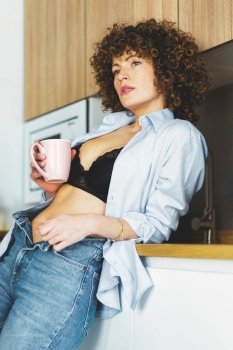 Curly haired female in black bra unbuttoned shirt and unzipped jeans leaning on counter while enjoying hot beverage. Adult woman with unbuttoned shirt drinking coffee in kitchen