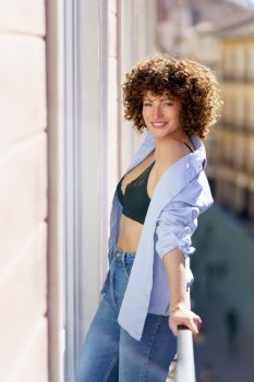 Adult positive female in unbuttoned shirt jeans and black bra standing on balcony against blurred background of town during weekend. Smiling curly haired woman in bra standing on balcony
