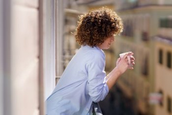 Side view of female with curly hair in blue shirt standing on small balcony and drinking hot beverage from cup against blurred background. Woman drinking hot tea on balcony against buildings