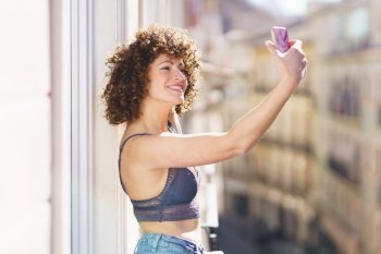 Cheerful young female in bra and jeans with curly hair taking selfie on smartphone while standing on balcony on sunny day. Smiling woman taking selfie on balcony