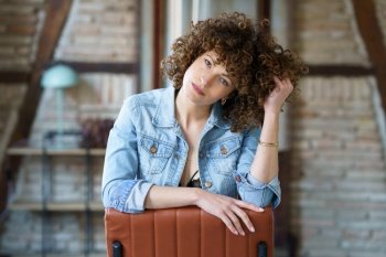 Serious young female in underwear and denim jacket sitting on leather chair and touching hair while looking at camera against blurred brick wall. Pensive woman sitting on chair and touching curly hair