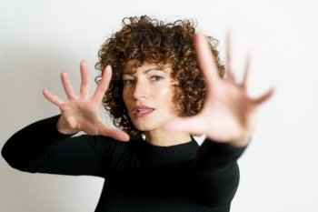 Attractive young female model, with curly hair looking at camera and showing stop gesture with full open palms and fingers against gray background in blurring unseen side illuminating lights. Focused woman stretching arms and showing stop gesture with full open palms