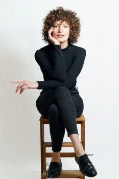 Concentrated and confident young female, in black outfit with hand touching cheek sitting on chair with legs crossed while leaning on elbow and looking at camera in gray backdrop. Focused curly haired woman sitting on wooden chair