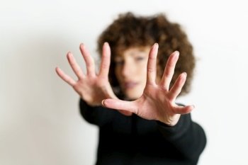 Defocused face of girl model, with curly hair looking at camera and showing stop gesture with full open palms and fingers against gray background in blurring unseen side illuminating lights. Defocused woman stretching arms and showing stop gesture with full open palms