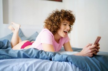 Full body side view of cheerful young female with curly dark hair lying on bed and browsing mobile phone during weekend at home. Happy woman using smartphone in bed at home