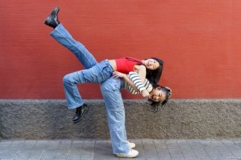 Side view of cheerful Asian woman in casual clothes lifting smiling friend on back while standing against red wall. Happy Asian female lifting friend on back