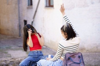 Young female photographer taking photo of girlfriend showing peace gesture on city street during vacation. Woman taking photo of girlfriend on camera on street