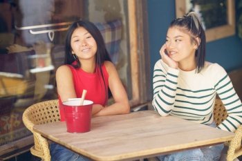 Smiling young Asian female friends in casual clothing sitting together at wooden table and discussing plans while spending time in cafe. Happy Asian women chatting in cafe together