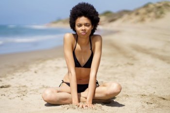 Full body of slim African American female traveler, in swimwear looking at camera with serious face, while sitting on sandy coast near sea during summer vacation. Serious black woman with Afro hair in bikini near sea