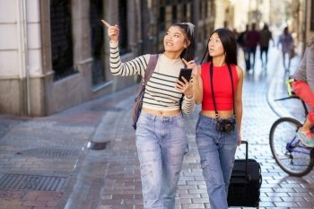 Young positive Asian girlfriends with backpack and suitcase walking on street of Granada and pointing at sightseeing while using cellphone during journey. Asian female travelers pointing away while walking on street