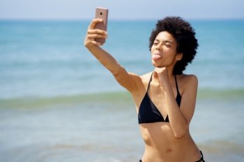 Funny African American female traveler in swimwear showing tongue and taking self portrait on smartphone while standing on coast near sea on sunny day. Happy black woman taking selfie on seashore in summer