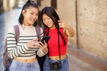 Asian girls in their 20s traveling through city of Granada using map on cellphone and pointing away while looking for direction during trip together. Asian girlfriends using map on smartphone while traveling together