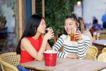 Cheerful young Asian female friends in casual wear sitting at wooden table and chatting while drinking refreshing cold tea during meeting in cafe. Smiling Asian friends drinking fresh beverages in cafe