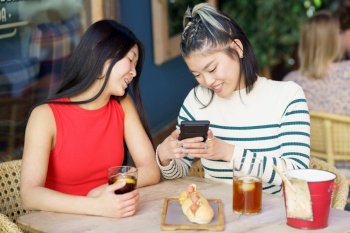 Content young Asian female friends in casual outfits, smiling and taking photo of delicious meal on smartphone while gathering at wooden table with glasses of drinks in cafe. Positive Asian women taking picture of yummy food in cafe