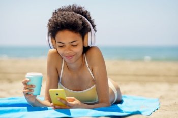 Content young ethnic female traveler with dark Afro hair, in stylish bikini and headphones, drinking takeaway coffee and messaging on smartphone while listening to music and sunbathing on sandy beach. Happy black lady using smartphone and drinking takeaway beverage lying on seashore