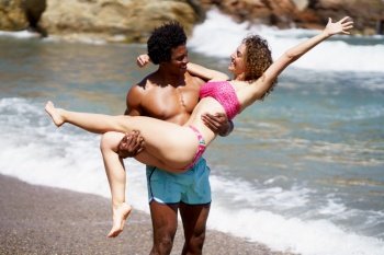 Smiling young multiracial couple enjoying near waving sea, and looking at each other while black male carrying girlfriend with stretched hand and standing on beach in daylight. Diverse couple having fun on beach during summer