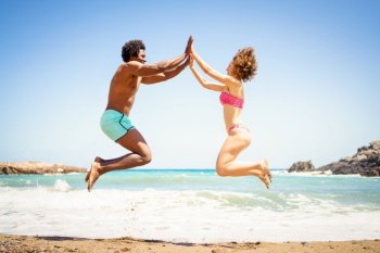 Full length side view of cheerful African American man and woman in bikini jumping on sandy seashore and holding hands while looking at each other. Happy multiethnic couple jumping near sea