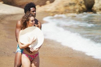 Young African American man embracing girlfriend from behind covering bare breast with hat while enjoying summer vacation near waving sea. Multiracial couple hugging on beach