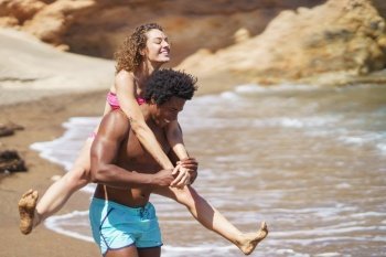 Cheerful young African American man giving piggyback ride to curly haired woman in bikini having fun on sandy beach near waving sea. Happy black man giving piggyback ride to girlfriend on beach