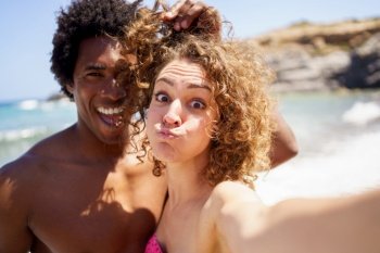 Cheerful young woman with puffed out cheeks taking selfie with black boyfriend touching curly hair of girlfriend while having fun on beach. Selfie of funny diverse couple on seashore