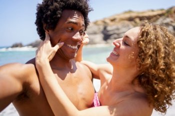 Cheerful young female with curly hair stretching corners of black boyfriends mouth making smile while taking selfie on beach. Selfie of cheerful woman stretching corners of boyfriends mouth
