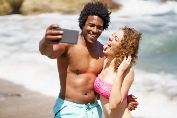 Happy African American man with naked torso and curly hair taking selfie on cellphone with girlfriend showing tongue against waving sea. Cheerful diverse couple taking selfie on seashore during sunny day