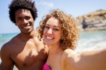 Cheerful multiracial girlfriend and boyfriend in swimwear smiling while taking selfie on sunny day near sea during summer holidays. Content diverse couple taking selfie on coast of sea
