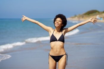 Positive African American female in swimwear with Afro hairstyle looking into distance with outstretched arms while standing on wet coast near sea. Gleeful black woman in bikini standing near sea