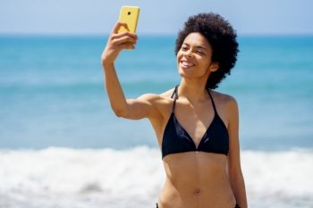 Joyful African American female traveler in bikini taking self portrait on cellphone with smile while standing near waving sea in tropical resort. Gleeful black woman in swimwear taking selfie on seashore