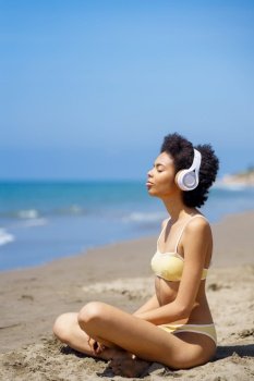 Side view of young fit ethnic female tourist with Afro hair in swimwear listening to music in headphones, while meditating on sandy beach with closed eyes in Lotus pose. Calm black woman listening to music with closed eyes while relaxing on seashore