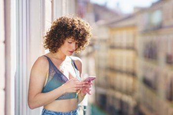 Positive young female in bra and jeans with curly hair while browsing smartphone on sunny balcony. Young female in bra and jeans with curly hair while browsing smartphone