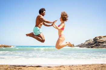 Full body side view of cheerful African American boyfriend and curly haired girlfriend, in swimwear jumping on sandy beach and holding hands near waving sea while looking at each other. Happy diverse couple jumping near sea